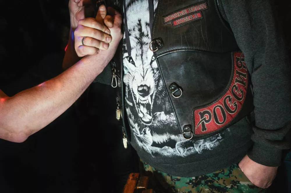 Deux personnes se serrent la main. L'une porte un gilet en cuir avec des inscriptions en cyrillique et un t-shirt avec une image de loup.