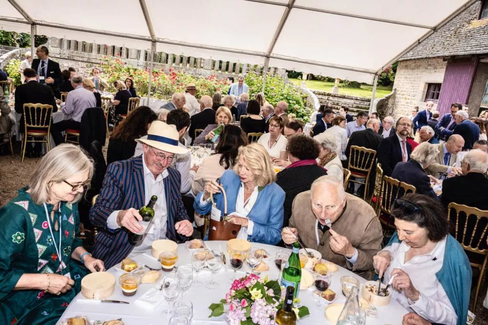 Un groupe de personnes est assis à une table sous une tente lors d'un événement en plein air. Ils sont en train de manger et de boire, entourés de nombreuses autres tables également occupées. L'ambiance semble conviviale et festive.