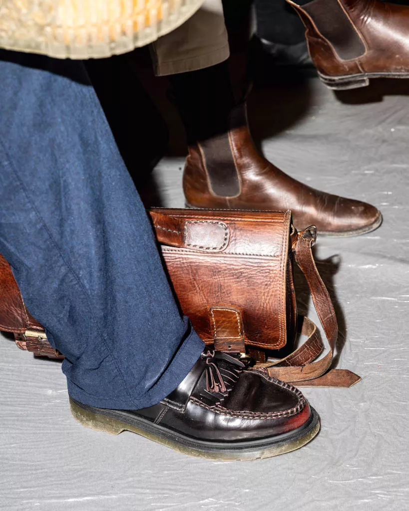 Des chaussures en cuir marron sont portées par des personnes, l'une portant des mocassins avec des glands et l'autre des bottines. Il y a également une sacoche en cuir marron posée au sol.