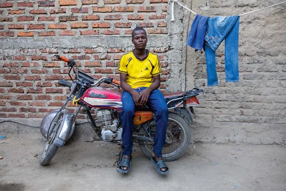 Un homme est assis sur une moto devant un mur en briques. Il porte un maillot jaune et un pantalon bleu. À côté, des vêtements sont suspendus sur une corde à linge.