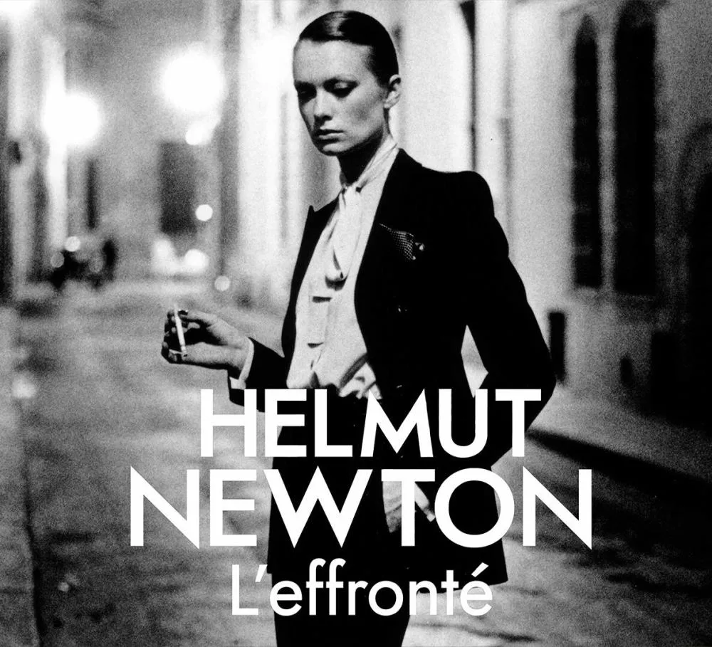 Une personne en costume élégant tient une cigarette dans une rue sombre. Le texte dit "Helmut Newton L'effronté".