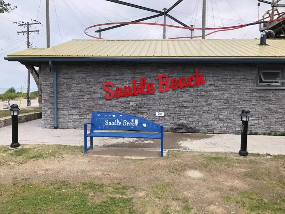Un bâtiment en pierre avec le nom "Sauble Beach" en lettres rouges sur le mur. Devant, un banc bleu avec le même nom écrit dessus.