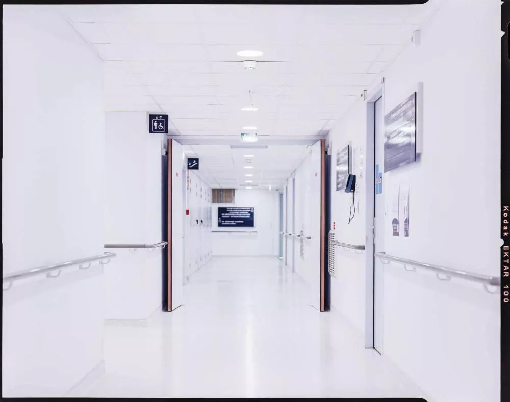Un couloir d'hôpital lumineux avec des murs blancs, des rampes de soutien, des portes fermées et des panneaux d'information.