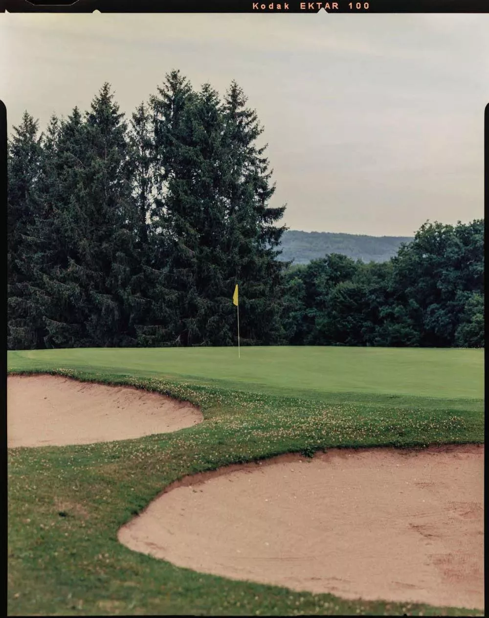Un terrain de golf avec des bunkers de sable au premier plan et un drapeau jaune sur le green, entouré d'arbres et de collines en arrière-plan.