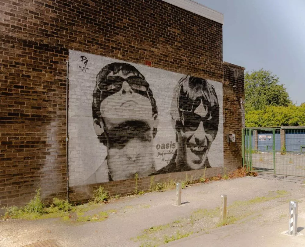 Une fresque murale en noir et blanc représentant deux personnes portant des lunettes de soleil, avec le mot "Oasis" et une signature en dessous.