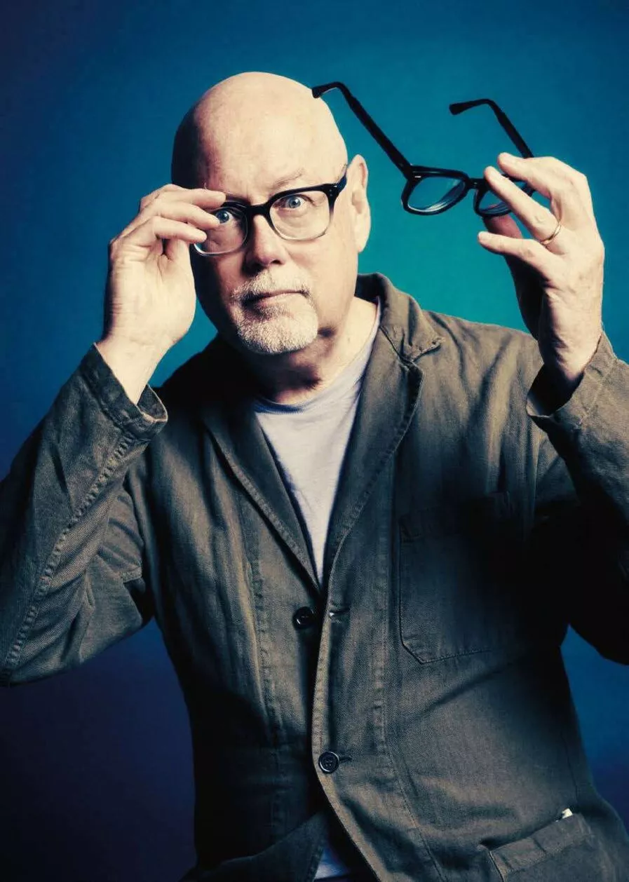 Un homme chauve avec une barbe blanche porte une veste et tient une paire de lunettes devant un fond bleu.
