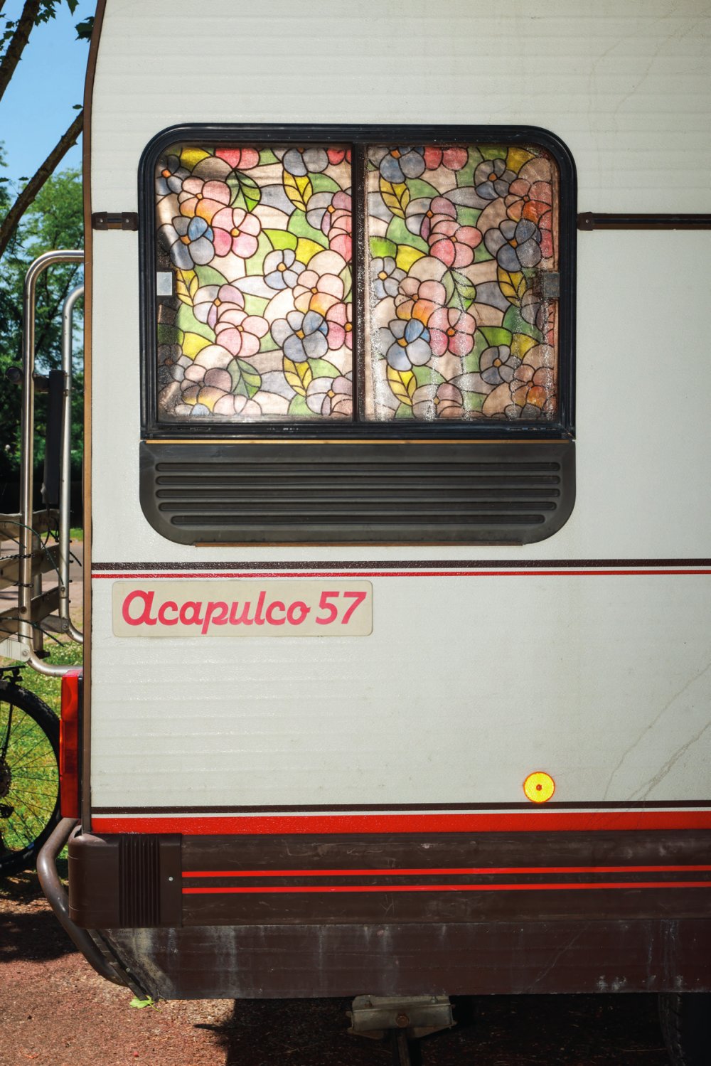 Une caravane avec une fenêtre décorée de motifs floraux colorés et le texte "Acapulco 57" écrit sur le côté.