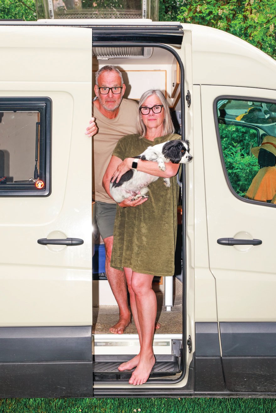 Un couple se tient à l'entrée d'un camping-car, l'homme derrière la femme qui tient un petit chien dans ses bras. Ils sont tous deux pieds nus et regardent vers l'extérieur.