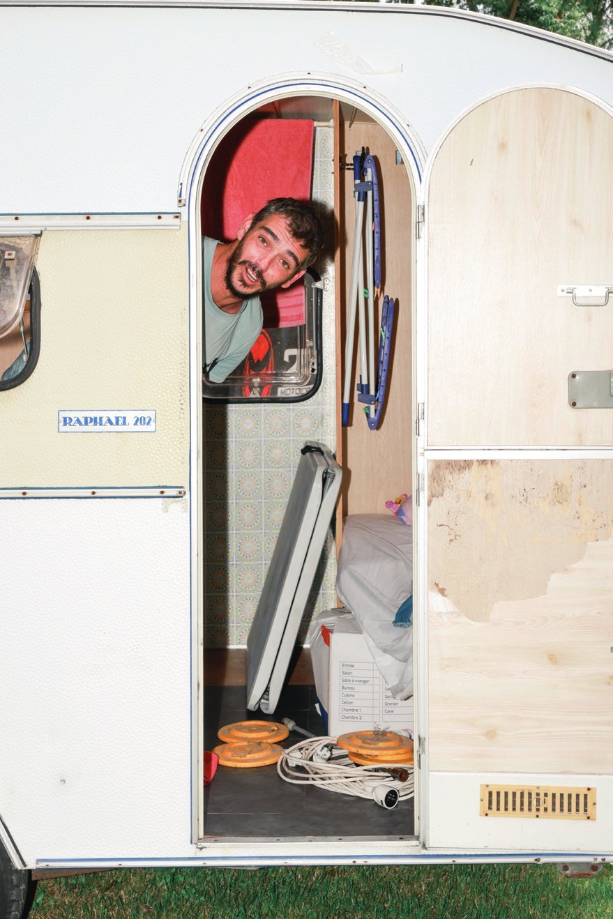 Un homme se penche par la porte ouverte d'une caravane, souriant. À l'intérieur, on peut voir des objets comme un matelas, des poids et des équipements de camping. La caravane est de couleur claire avec un panneau portant l'inscription "RAPHAEL 202".
