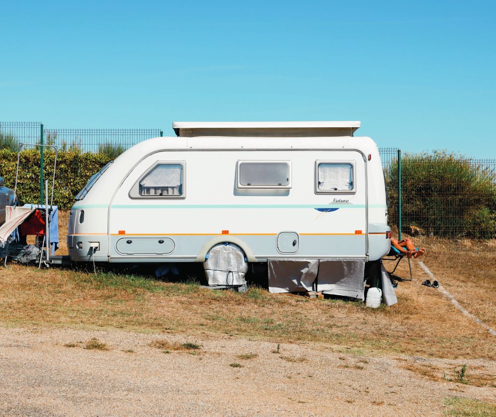 Une caravane blanche est stationnée sur un terrain herbeux, avec des buissons et une clôture en arrière-plan. Une personne est assise près de la caravane, avec les jambes visibles. Le ciel est bleu et dégagé.