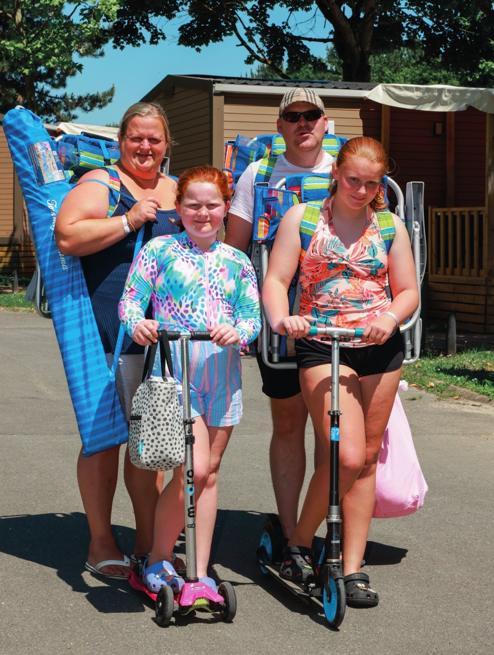Une famille de quatre personnes est en extérieur, portant des chaises de plage et des sacs, et deux enfants sont sur des trottinettes. Ils semblent prêts pour une journée à la plage ou en plein air.