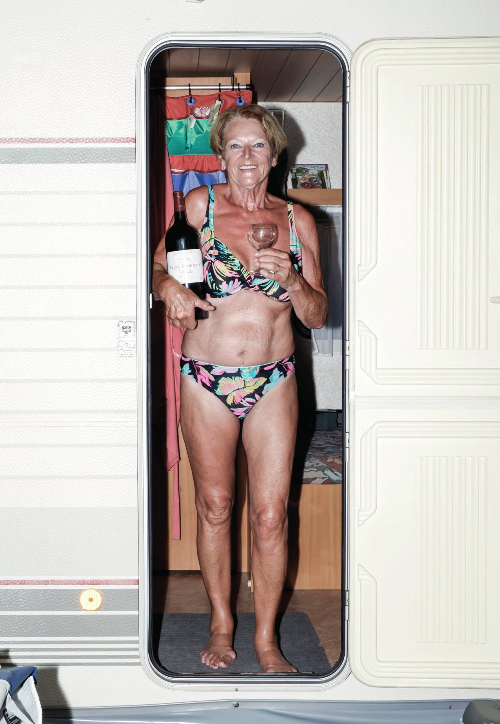 Une femme en maillot de bain à motifs floraux se tient dans l'entrée d'une caravane, tenant une bouteille de vin et un verre.