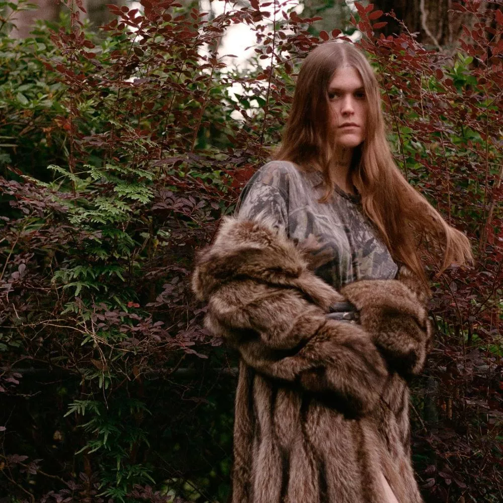 Une personne aux cheveux longs se tient dans un environnement forestier, portant un manteau en fourrure et un vêtement à motif camouflage.