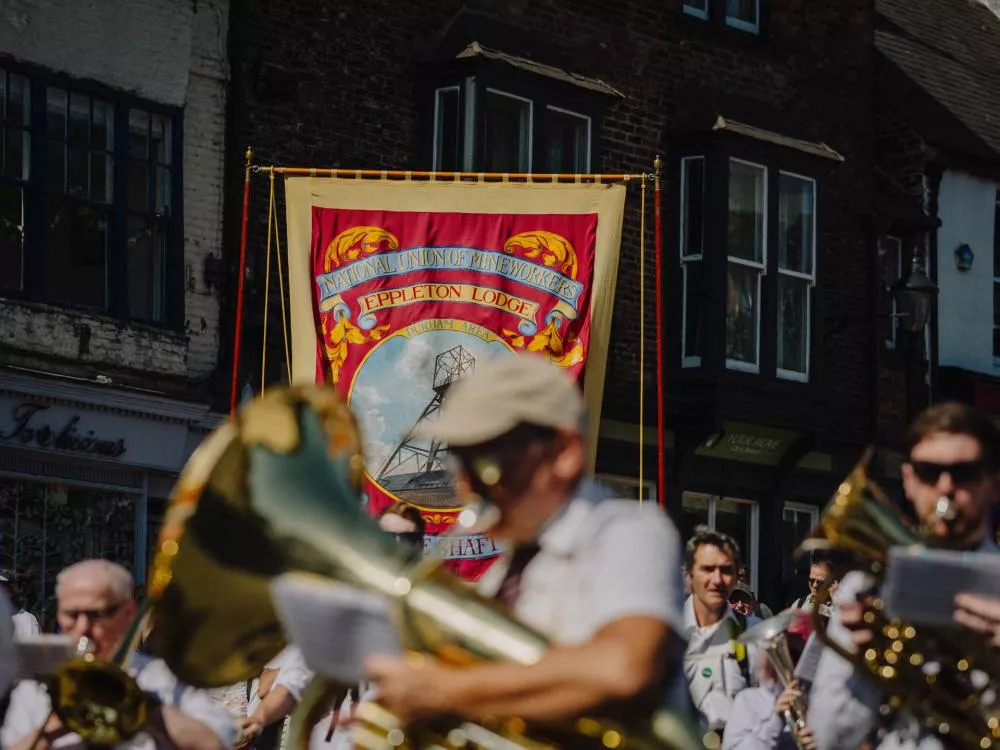 Un groupe de musiciens défile dans la rue avec des instruments à vent. En arrière-plan, une bannière colorée indique "National Union of Mineworkers, Eppleton Lodge, Durham Area" avec une image d'une structure minière.