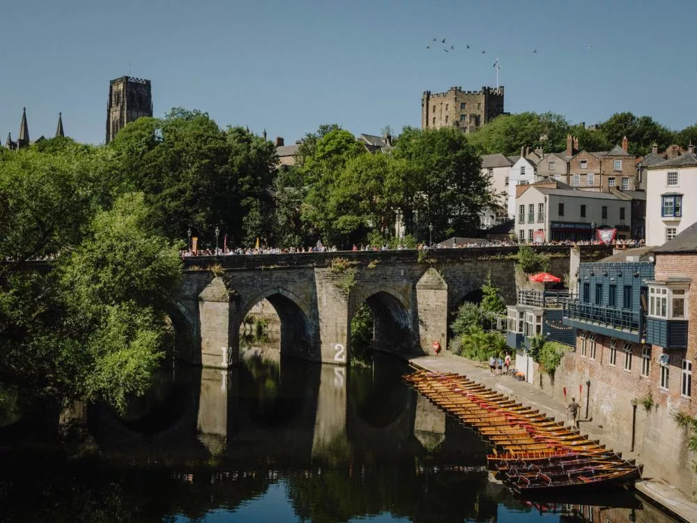 Une vue d'un pont en pierre avec des arches au-dessus d'une rivière, entouré de bâtiments historiques et d'arbres. Des barques sont alignées le long de la rive, et une foule de personnes se trouve sur le pont. On aperçoit également une cathédrale et un château en arrière-plan.