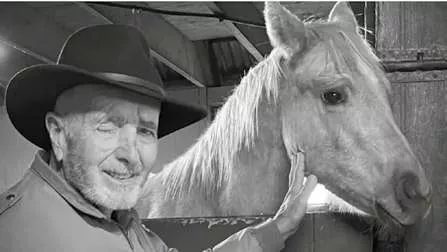 Un homme portant un chapeau caresse un cheval dans une écurie. L'image est en noir et blanc.