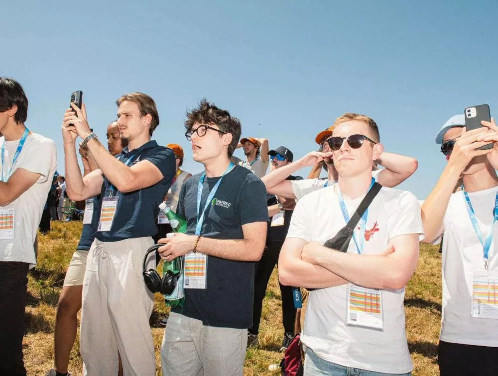 Un groupe de personnes, principalement des hommes, se tient à l'extérieur par une journée ensoleillée. Certains prennent des photos avec leurs téléphones, d'autres portent des lunettes de soleil et des badges autour du cou. Ils semblent assister à un événement.