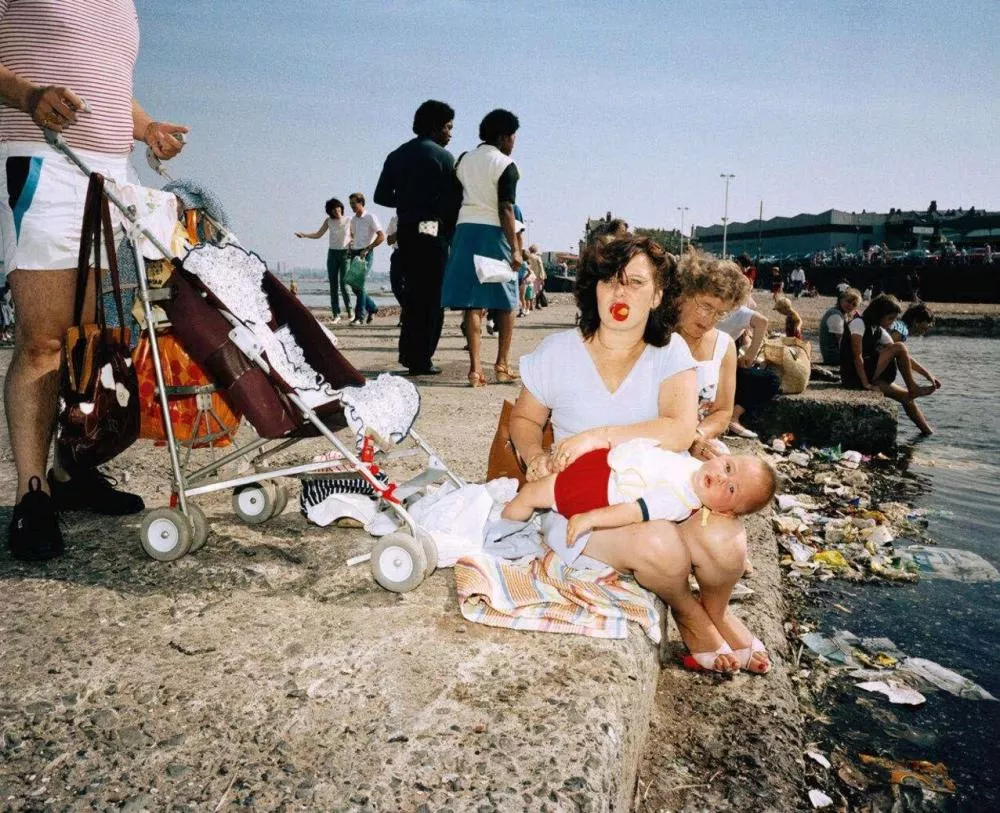 Scène de bord de mer avec plusieurs personnes, dont une femme assise tenant un bébé sur ses genoux. Un landau est visible à côté. L'environnement est animé avec des gens se promenant et d'autres assis près de l'eau.