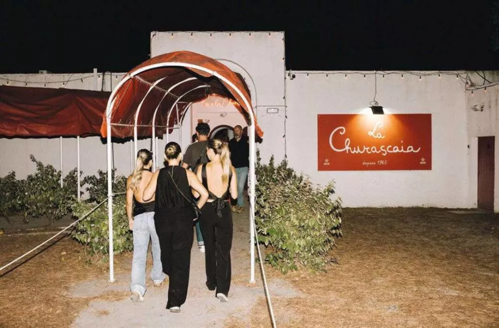 Un groupe de personnes entre dans un bâtiment sous un auvent rouge. Sur le mur à droite, il y a une enseigne rouge indiquant "La Churrascaia depuis 1965".