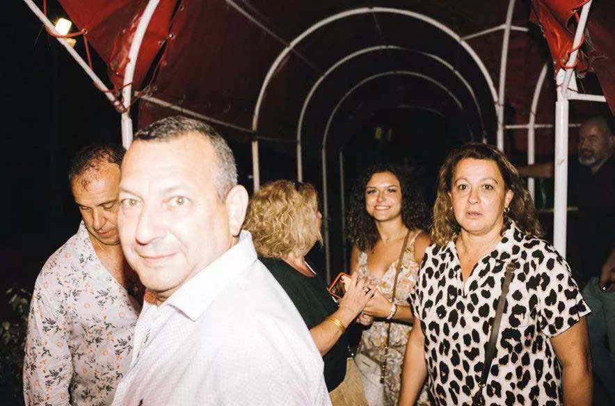 Un groupe de personnes se tient sous un auvent rouge. Certaines regardent vers l'objectif, d'autres discutent entre elles. L'ambiance semble être celle d'un événement social ou d'une fête.