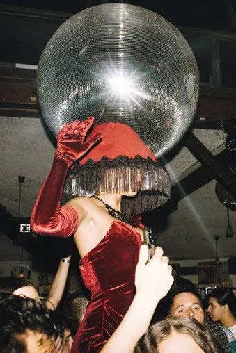 Une personne en robe rouge et gants assortis porte un grand abat-jour rouge sur la tête, entourée de gens dans ce qui semble être une fête ou un club, avec une boule disco en arrière-plan.