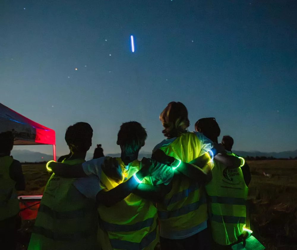 Un groupe de personnes portant des gilets réfléchissants et des bracelets lumineux regarde le ciel nocturne où l'on peut voir une traînée lumineuse. Une tente est visible sur la gauche.
