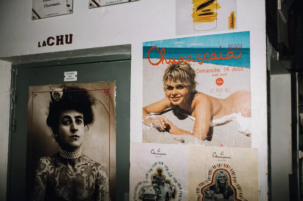 L'image montre un mur avec plusieurs affiches. Une affiche colorée montre une femme souriante sur une plage avec le texte "Churascaia Beach" et une date. Une autre affiche en noir et blanc représente une personne tatouée avec un collier de perles. Il y a aussi des petites affiches avec le texte "La Churascaia" et "Les Enfants de la Chu".