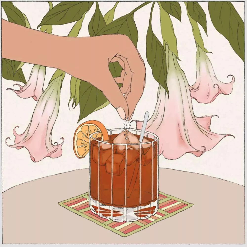 Une main saupoudre du sel dans un verre de boisson glacée avec une tranche de citron, sur une table avec un dessous de verre coloré, entourée de grandes fleurs roses retombantes.