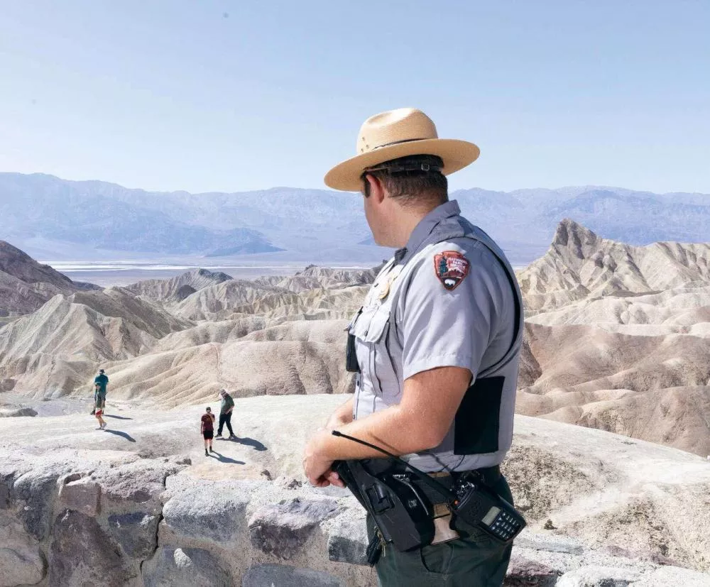Un garde forestier en uniforme, portant un chapeau, se tient sur un point de vue rocheux et regarde trois personnes marchant dans un paysage désertique montagneux.