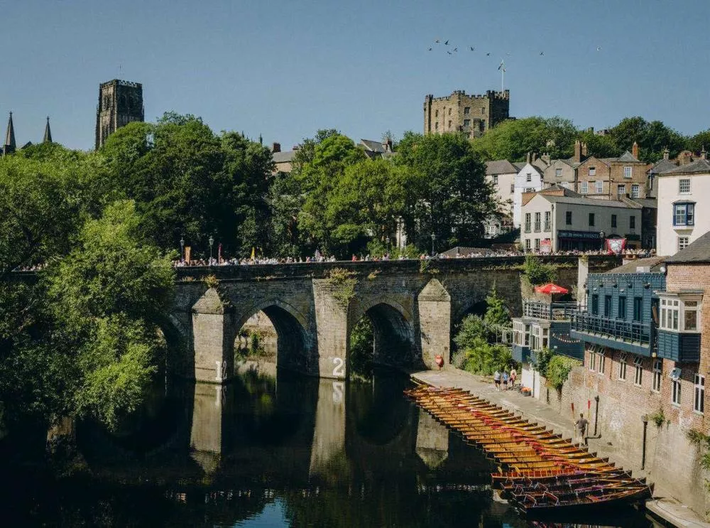 Un pont en pierre traverse une rivière, avec des barques alignées sur la rive. En arrière-plan, on voit des bâtiments historiques entourés d'arbres, et un château surplombe la scène. Des personnes se promènent sur le pont sous un ciel dégagé.