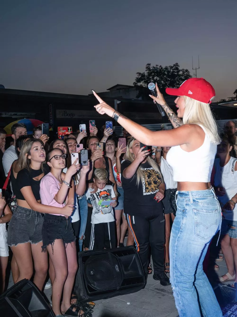 Une femme en tenue décontractée, portant une casquette rouge, semble animer un événement en plein air devant un public enthousiaste qui prend des photos.
