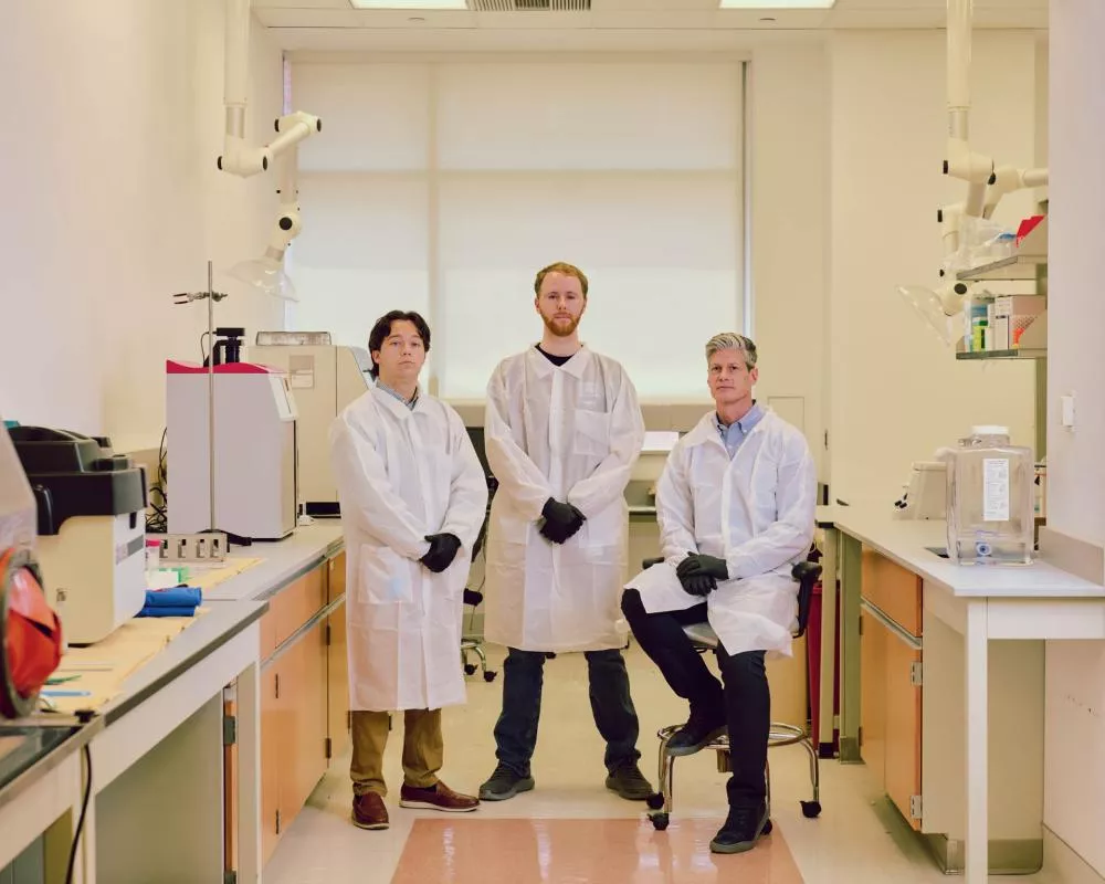 Trois personnes en blouses de laboratoire et gants noirs se tiennent dans un laboratoire équipé de divers appareils scientifiques. L'une est assise sur un tabouret tandis que les deux autres se tiennent debout.