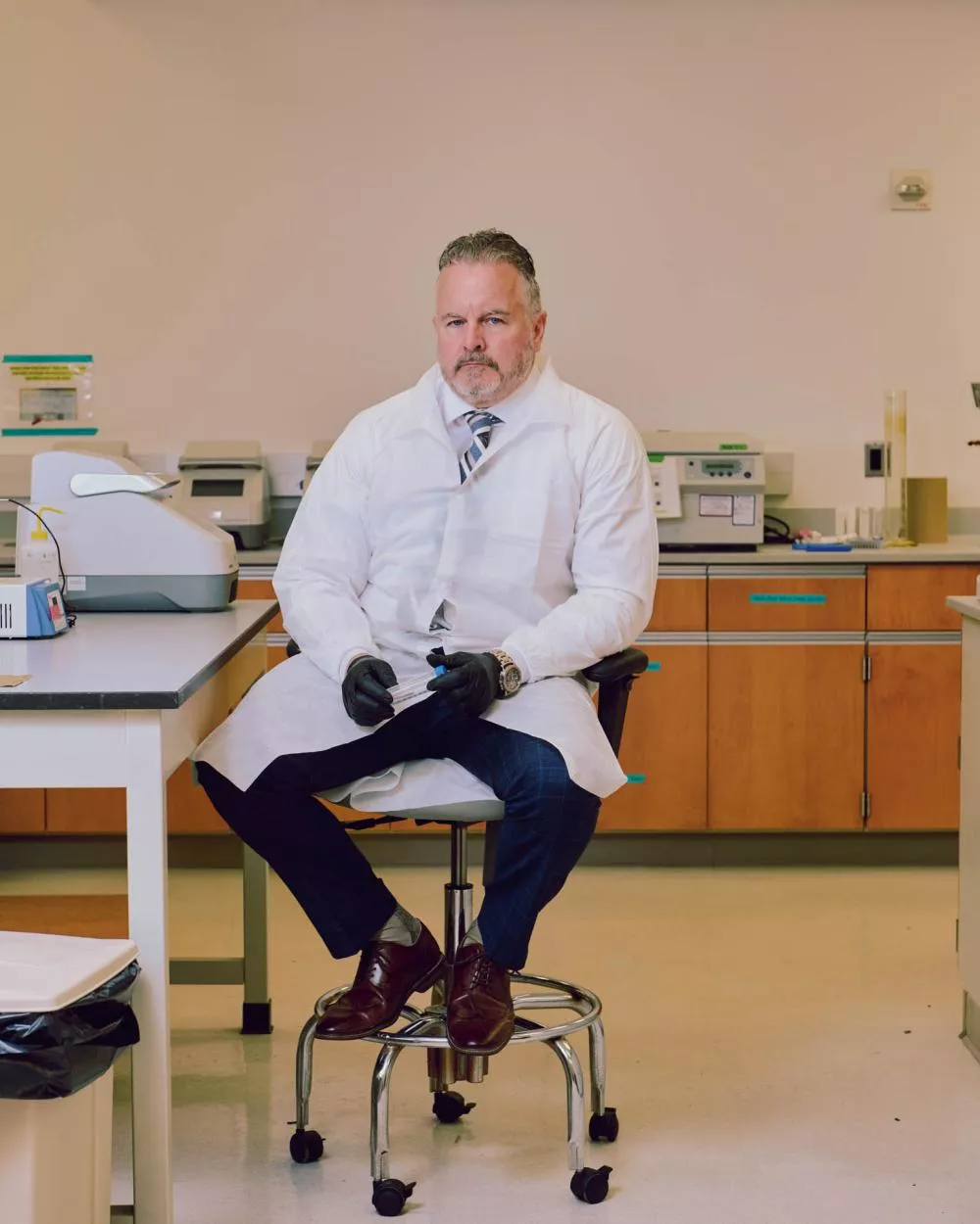 Un homme en blouse de laboratoire est assis sur un tabouret dans un laboratoire. Il porte des gants noirs et un costume avec une cravate. Autour de lui, il y a divers équipements de laboratoire sur les tables.
