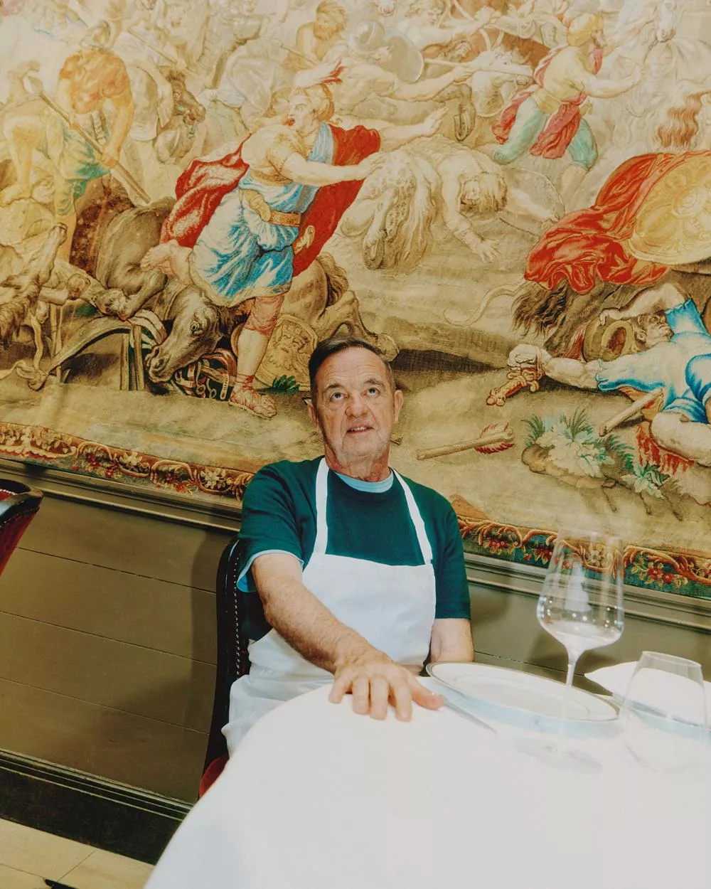 Un homme portant un tablier blanc est assis à une table de restaurant, devant une tapisserie colorée représentant une scène historique ou mythologique. Sur la table, il y a des verres à vin et une assiette vide.