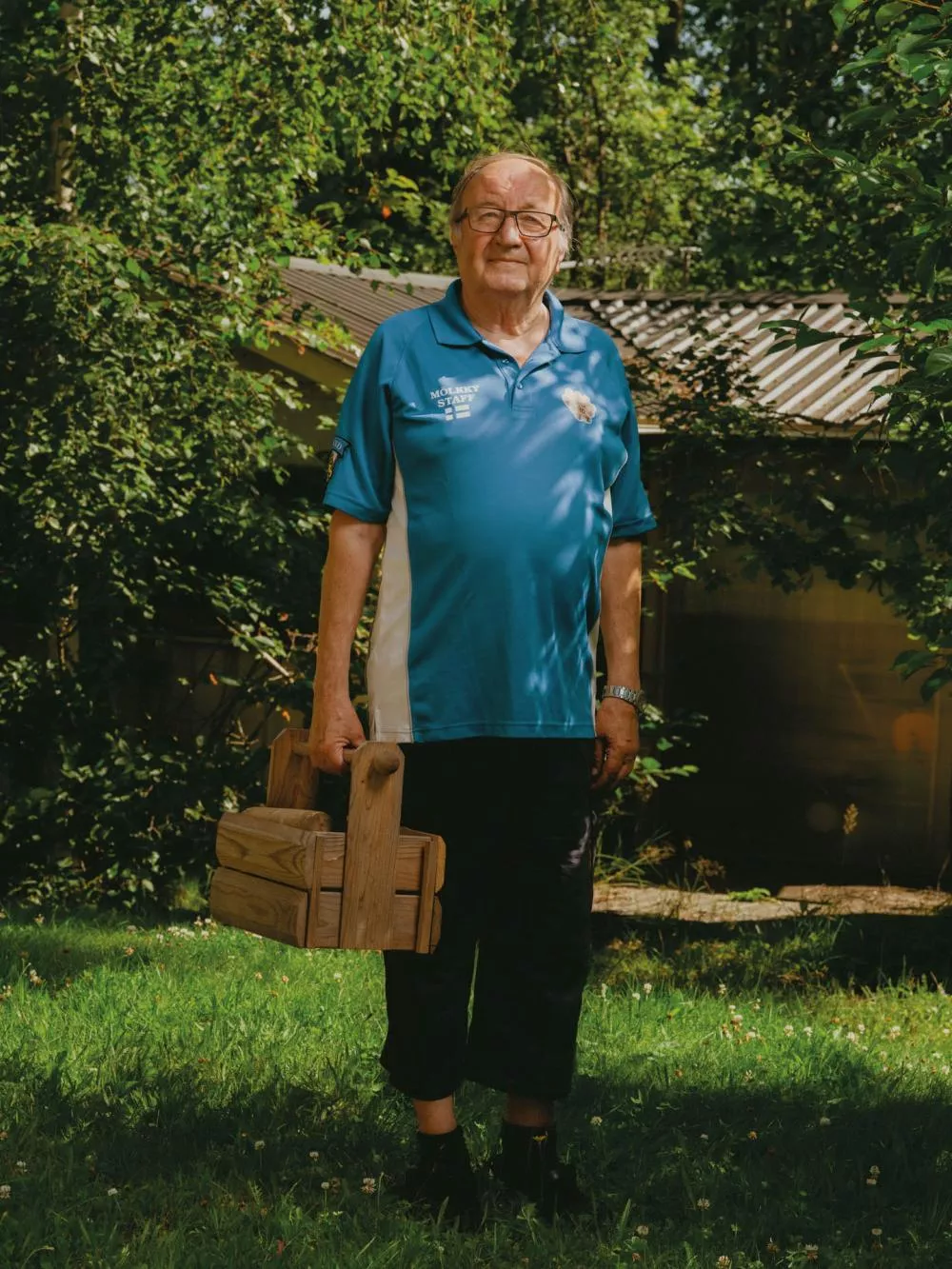 Un homme se tient debout dans un jardin verdoyant, portant un maillot bleu avec l'inscription "Mölkky Staff". Il tient une caisse en bois à la main.