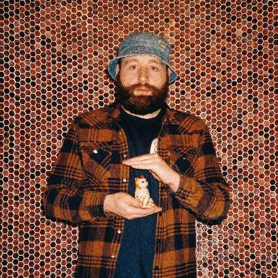 Un homme portant une chemise à carreaux et un bob tient une petite figurine de chat devant un mur recouvert de motifs hexagonaux.