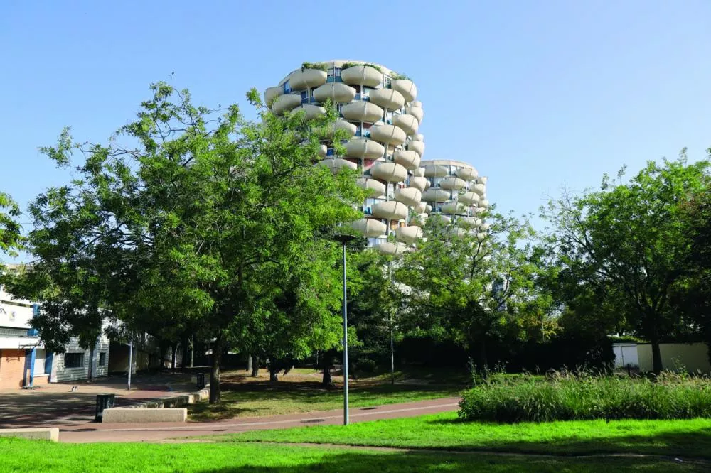 Un paysage urbain avec des bâtiments circulaires en arrière-plan, entourés de verdure et d'arbres sous un ciel bleu.