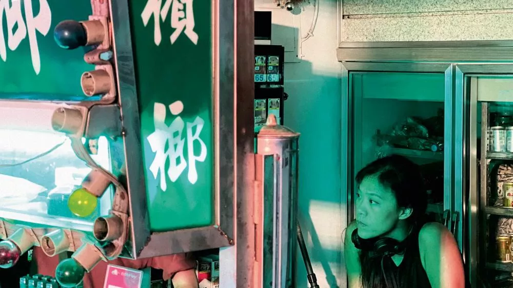 Une femme avec un casque autour du cou se tient près d'un stand ou d'un magasin éclairé par des lumières colorées, avec des inscriptions en caractères asiatiques sur un panneau vert.