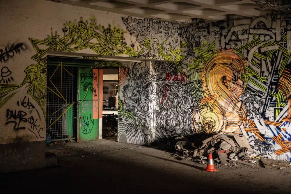 Un mur intérieur est couvert de graffitis colorés et complexes, avec des motifs géométriques et abstraits en jaune, orange et noir. Il y a une porte verte ouverte et des débris au sol, accompagnés d'un cône de signalisation orange.