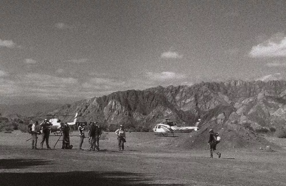 Un groupe de personnes se tient près d'un hélicoptère sur un terrain dégagé, avec des montagnes en arrière-plan. L'image est en noir et blanc et semble granuleuse.