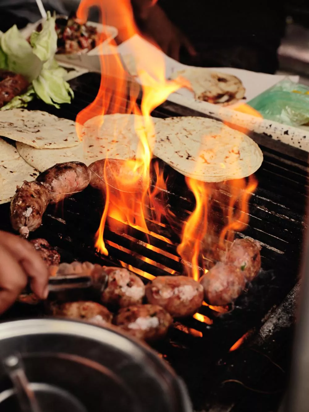 Des saucisses et des tortillas sont en train de cuire sur un gril avec des flammes vives. On voit également des légumes et des pinces pour manipuler les aliments.
