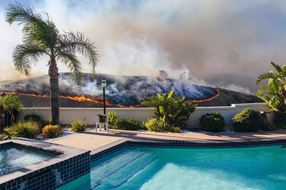 Un feu de végétation brûle sur une colline derrière une maison avec une piscine et un jardin bien entretenu.