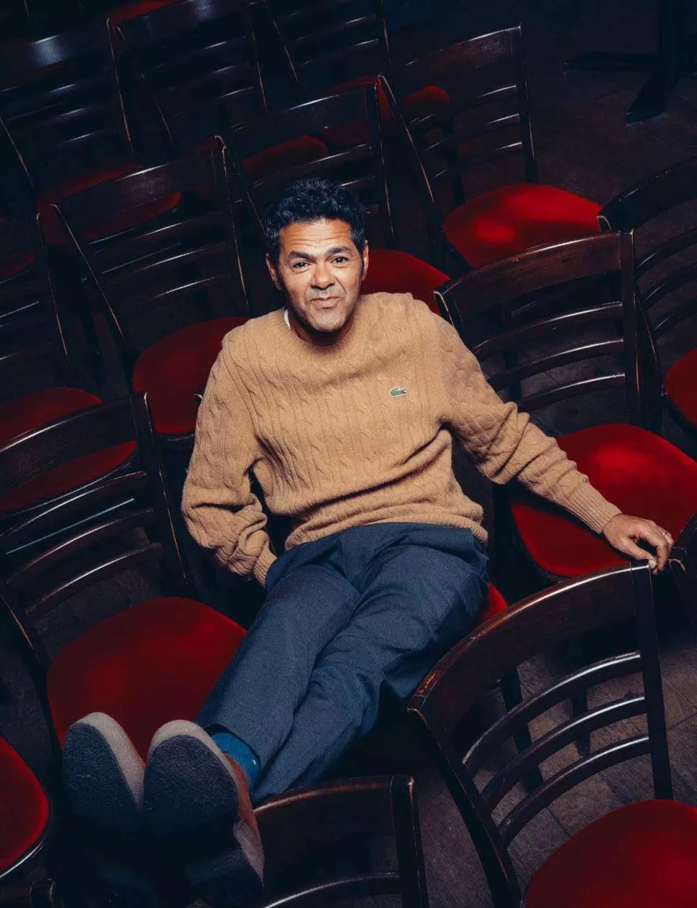 Un homme est assis confortablement sur une chaise au milieu de plusieurs autres chaises avec des sièges rouges. Il porte un pull beige et un pantalon bleu.