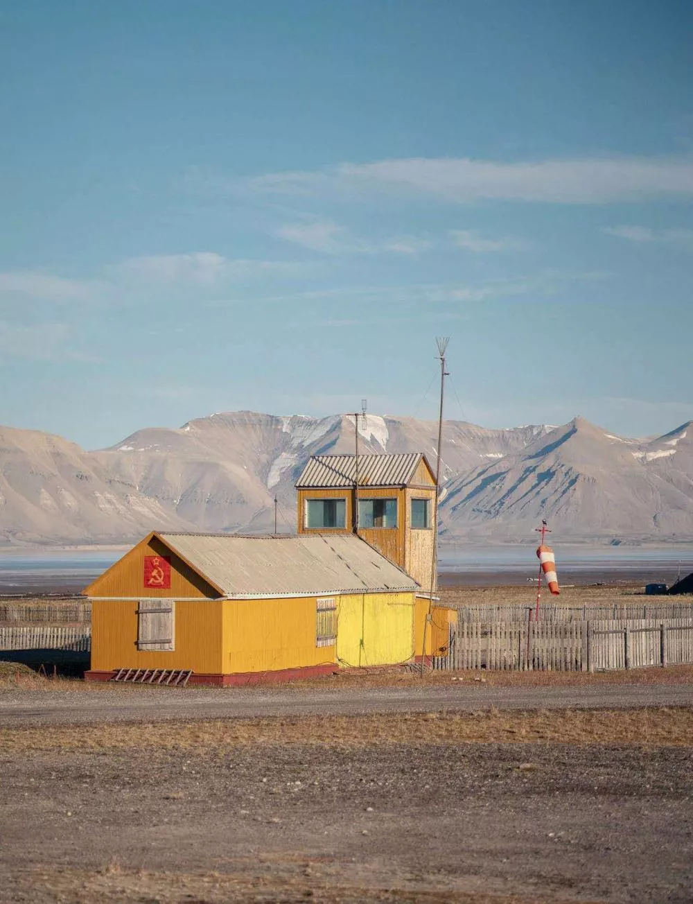 Un bâtiment en bois jaune avec un toit en tôle, situé dans un paysage montagneux aride. Une manche à air est visible à droite. Le bâtiment arbore un symbole rouge avec une faucille et un marteau.