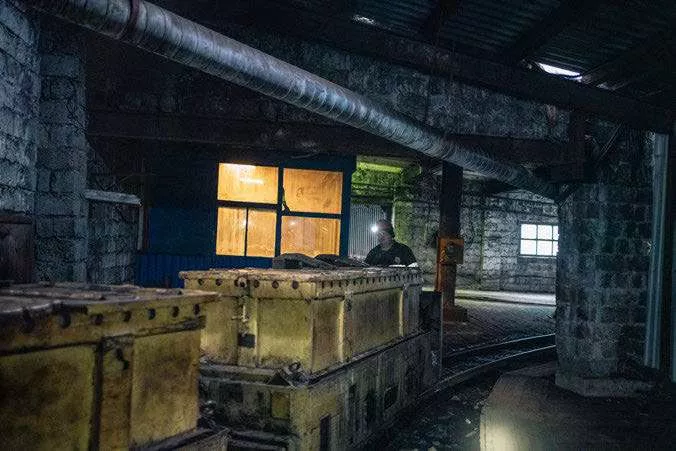 Un homme se tient à côté de wagons de mine dans un environnement industriel sombre, éclairé par une faible lumière provenant d'une fenêtre.