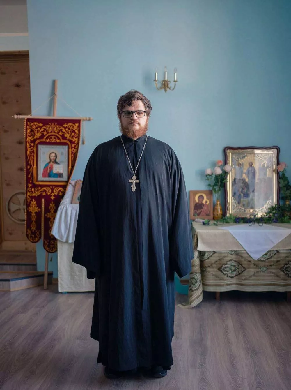 Un homme en habit religieux orthodoxe se tient debout dans une pièce ornée d'icônes et de décorations religieuses.