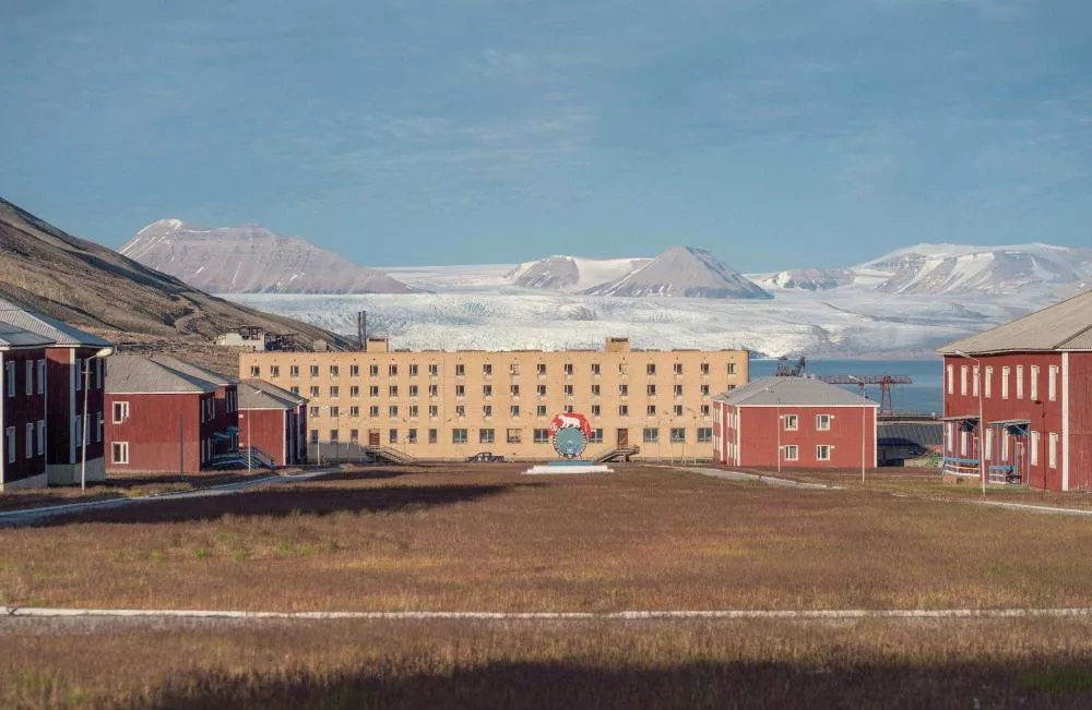 Un bâtiment central en brique claire est entouré de plusieurs bâtiments rouges. À l'arrière-plan, on voit des montagnes enneigées et un glacier sous un ciel dégagé.