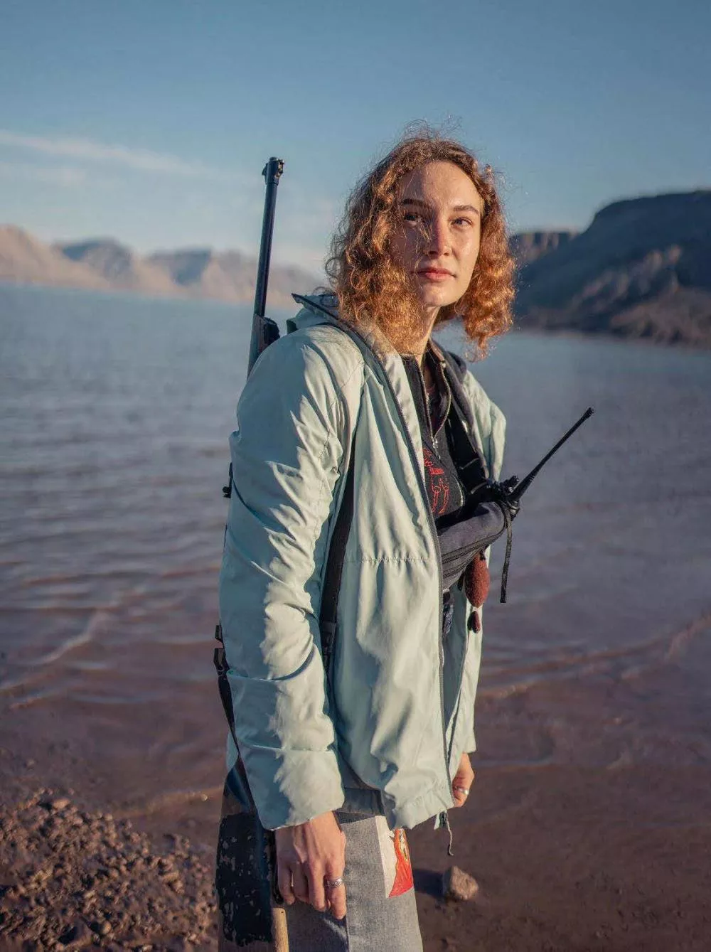 Une personne avec des cheveux bouclés porte une veste légère et tient un talkie-walkie. Elle se tient près d'un plan d'eau avec des montagnes en arrière-plan.