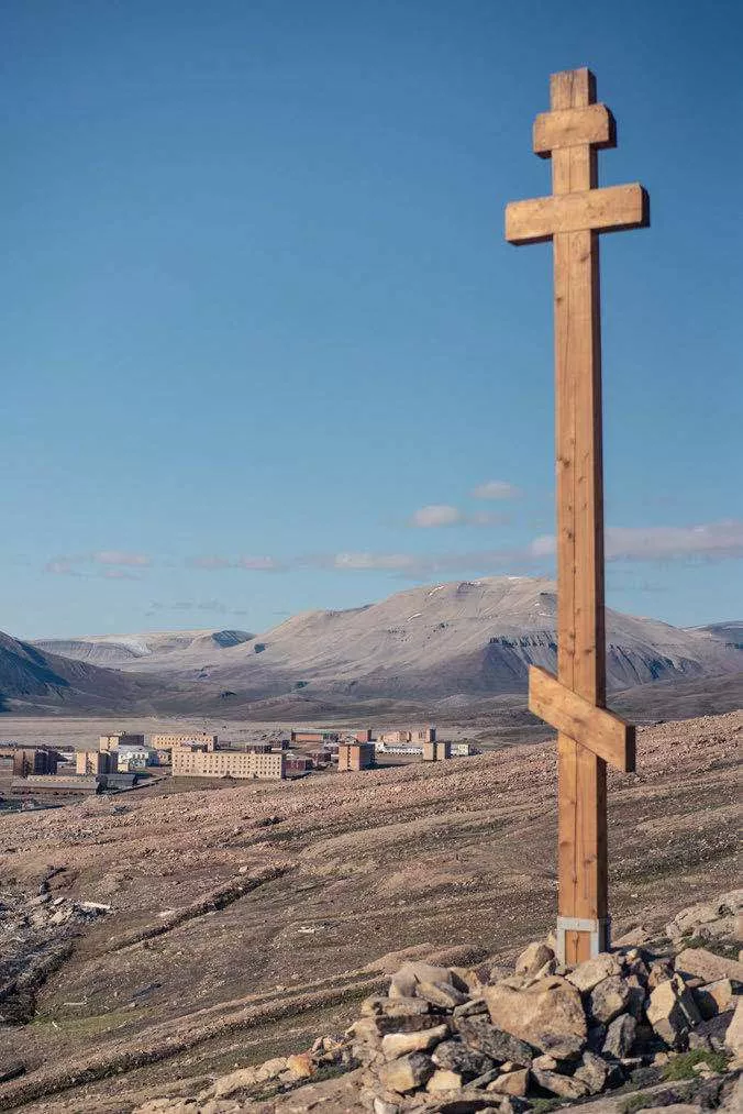 Une croix orthodoxe en bois est plantée sur un monticule de pierres au premier plan, avec en arrière-plan un paysage montagneux et un petit village ou complexe de bâtiments sous un ciel bleu.
