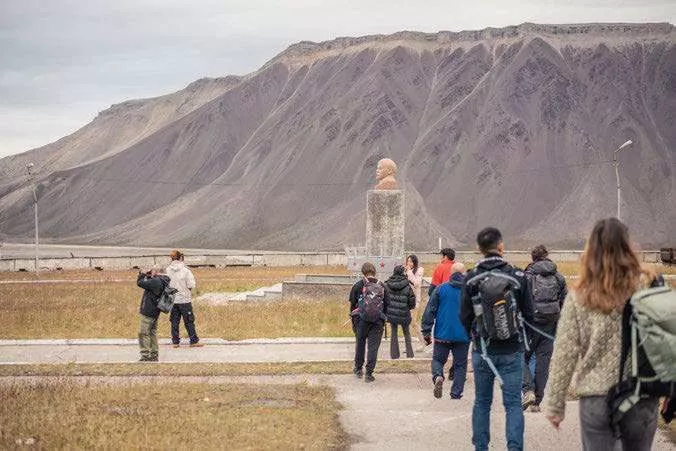 Un groupe de personnes marche vers une statue située devant une montagne. La statue semble être un buste posé sur un socle. L'environnement est naturel avec de l'herbe et un ciel nuageux.